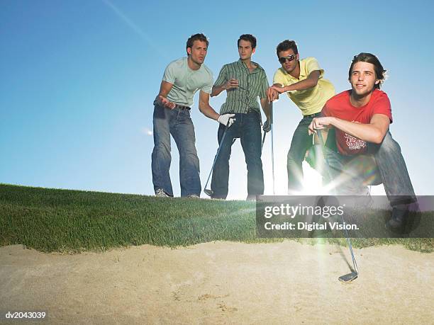 four young men at the edge of a sand bunker holding golf clubs - golf bunker low angle stock pictures, royalty-free photos & images