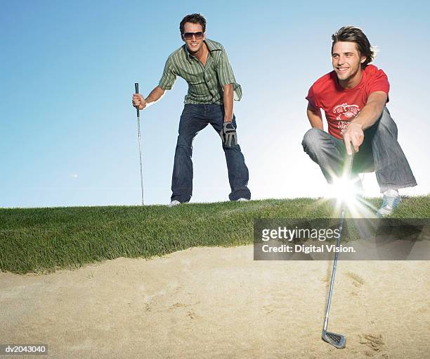 two young men standing at the edge of a sand bunker holding golf clubs - golf bunker low angle stock pictures, royalty-free photos & images