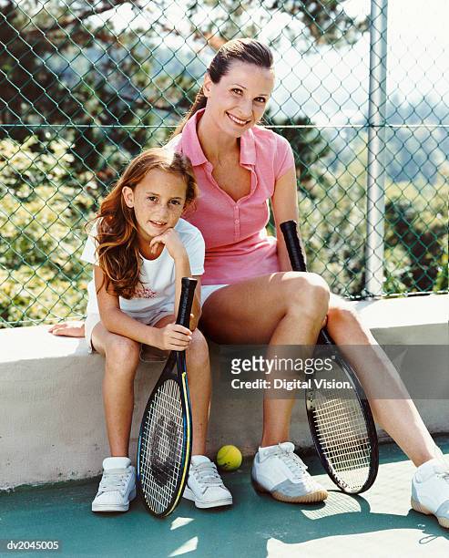 portrait of a woman and her young daughter sitting on the wall of a tennis court, holding racquets - tennis court stock pictures, royalty-free photos & images