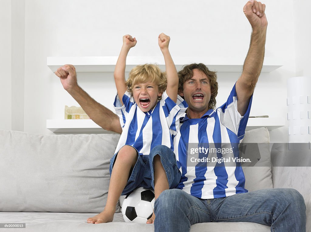 Father and Son Sitting on a Sofa, Wearing Football Strips and Cheering