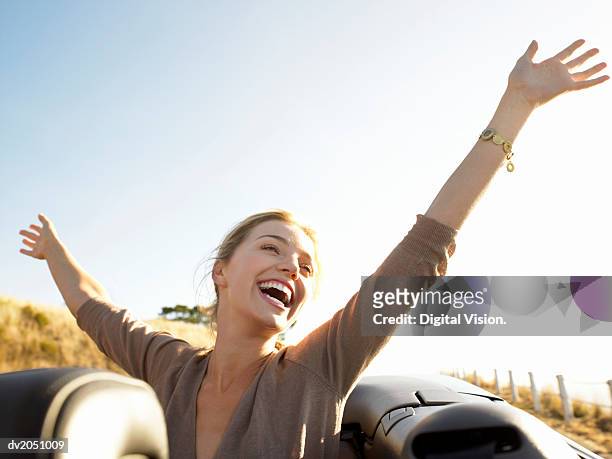 young woman sits in the back of a convertible, her arms in the air, laughing with joy - braccia alzate foto e immagini stock
