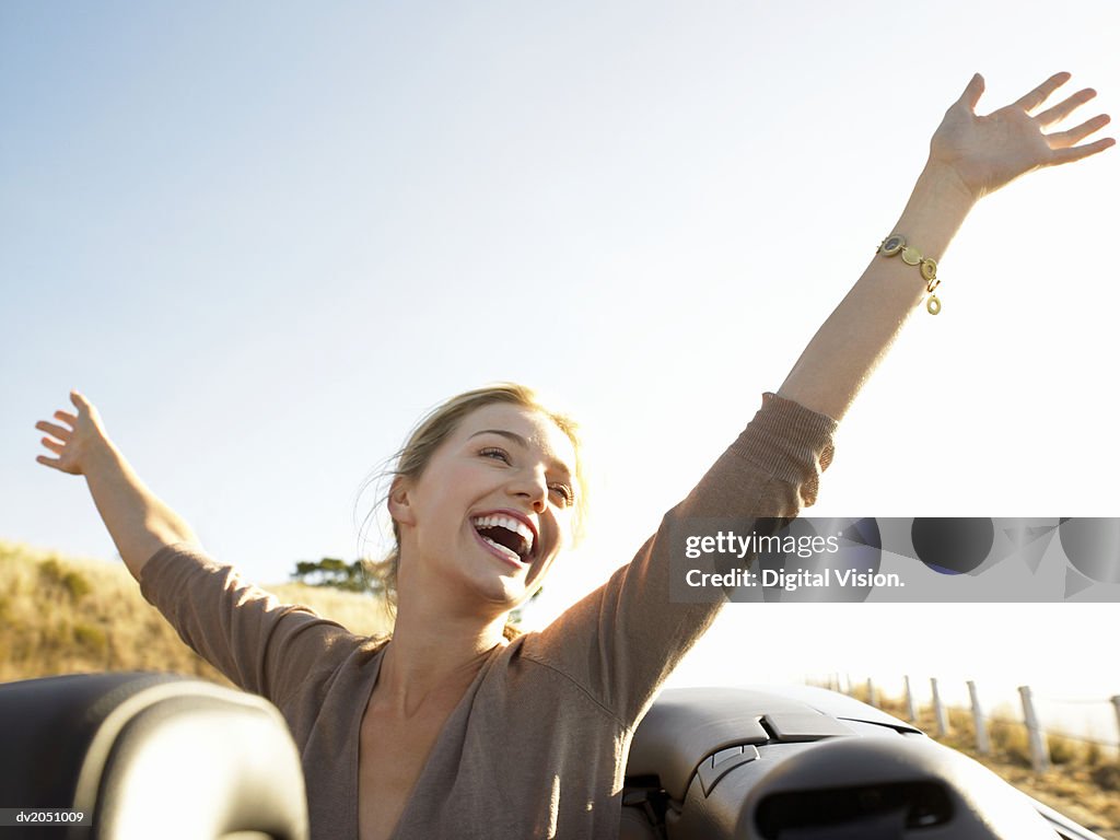 Young Woman Sits in the Back of a Convertible, Her Arms in the Air, Laughing With Joy