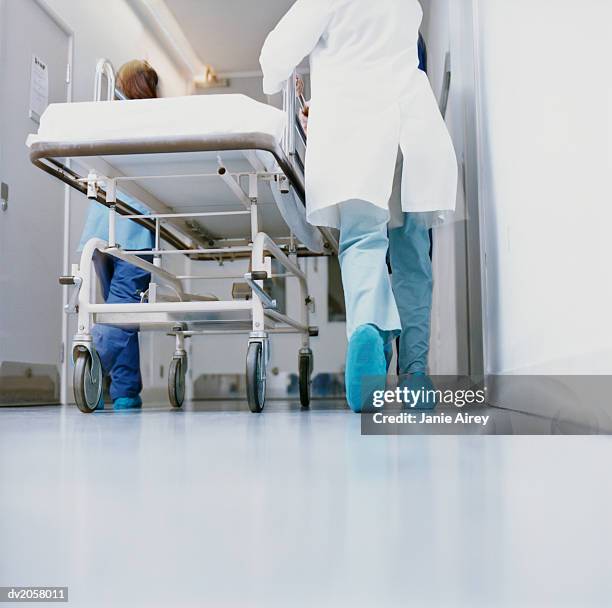 rear view of doctors in surgical scrubs hurrying down a hospital corridor with a trolley - hospital gurney stock pictures, royalty-free photos & images