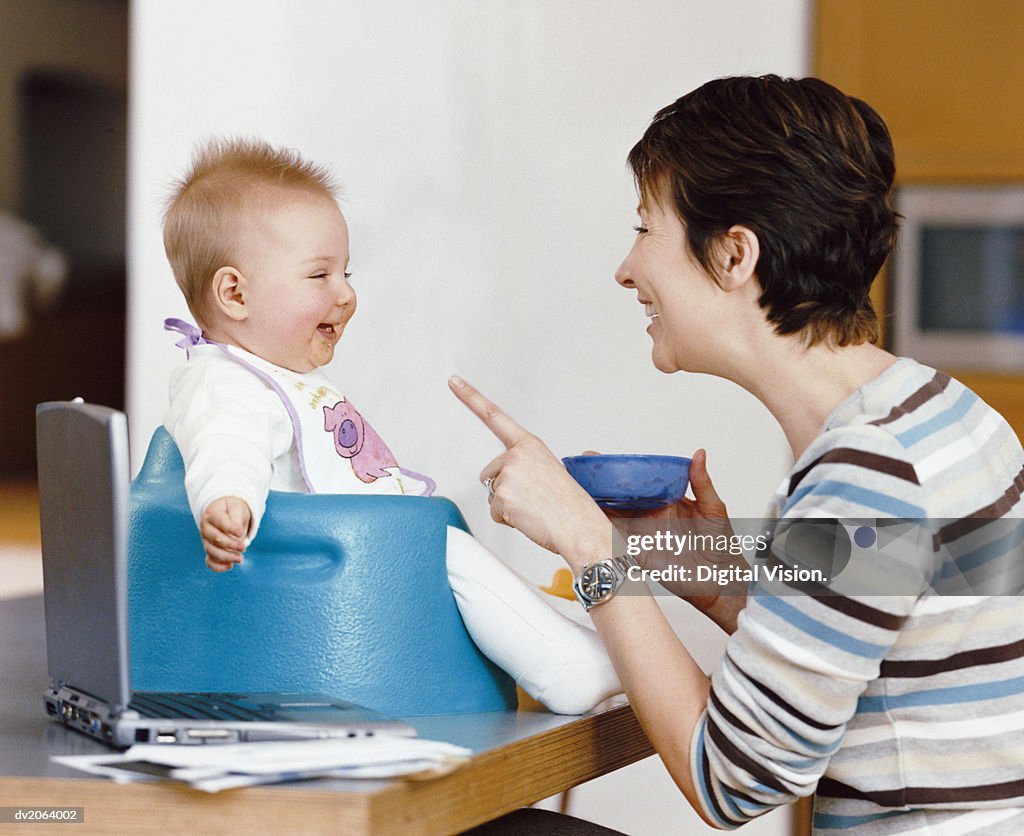 Mother Holds a Bowl of Baby Food, Talking and Smiling at Her Baby in a Highchair