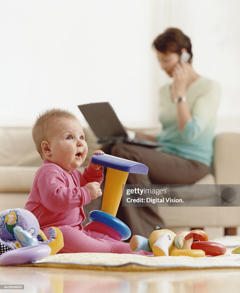 Baby Sits on the Floor Playing With Toys, the Mother in the Background