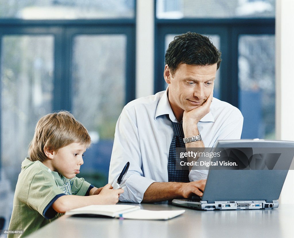 Businessman Using a Laptop and His Son Holding a Pen