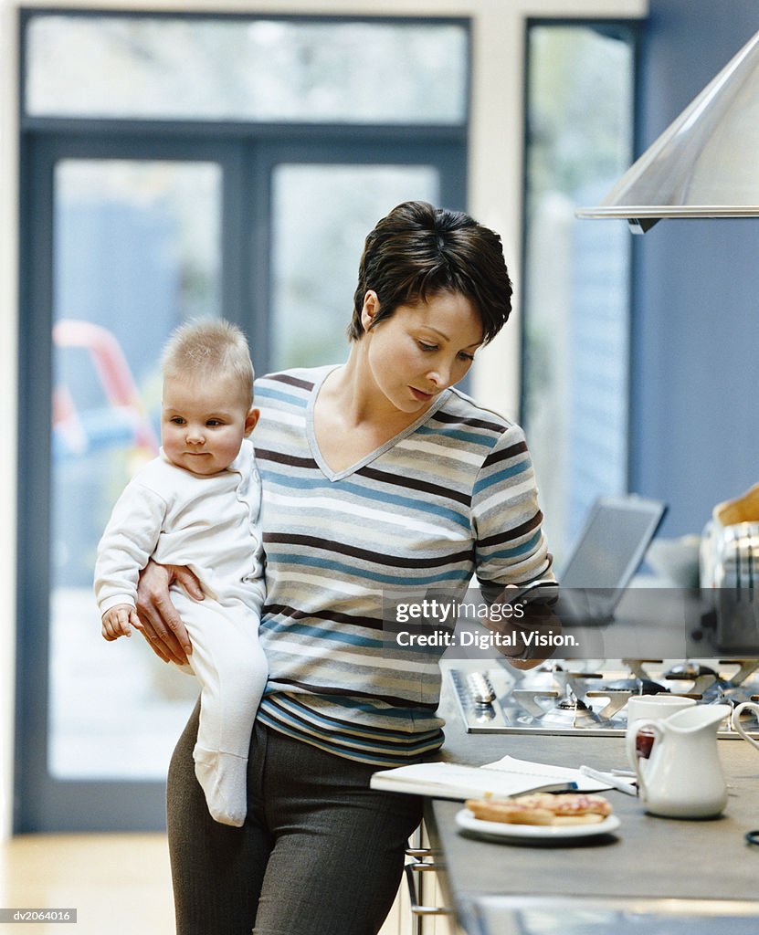 Woman Stands by a Kitchen Counter Holding Her Baby and Dialing Her Mobile Phone