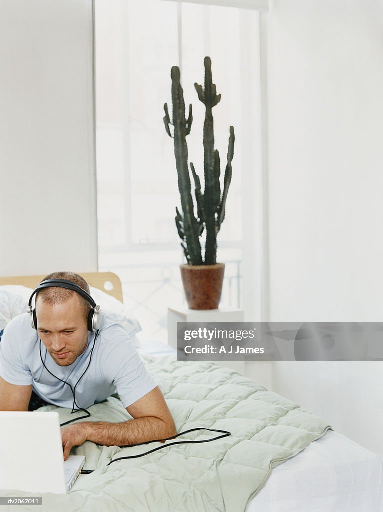 Man Lying on a Bed Using a Laptop Computer and Wearing Headphones