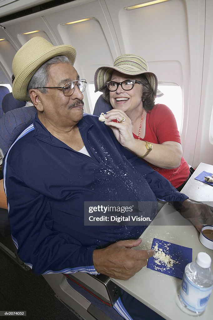 Senior Woman Feeding Biscuit to a Man Covered in Crumbs on a Passenger Aeroplane