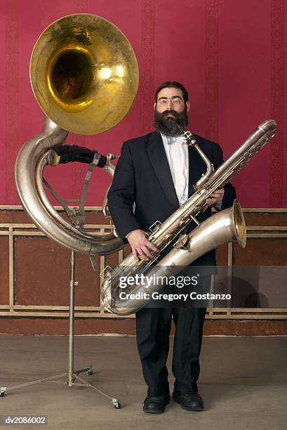 old fashioned man in a suit holding a large saxophone, standing next to a tuba on a music stand - greggory stock-fotos und bilder