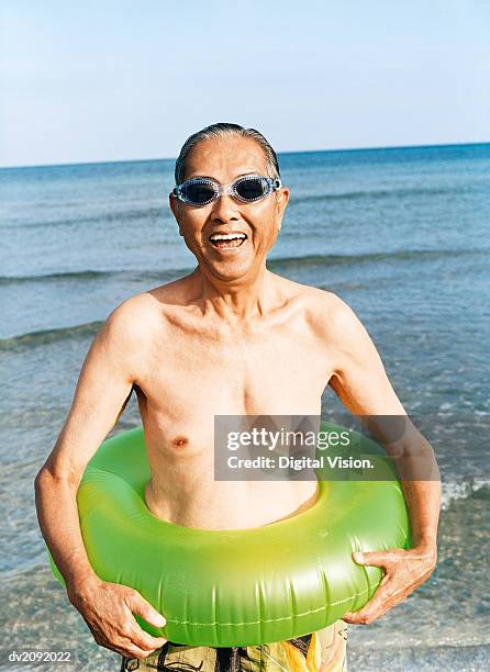 portrait of a smiling senior man standing by the sea, wearing a rubber ring and swimming goggles - rubber ring stock-fotos und bilder