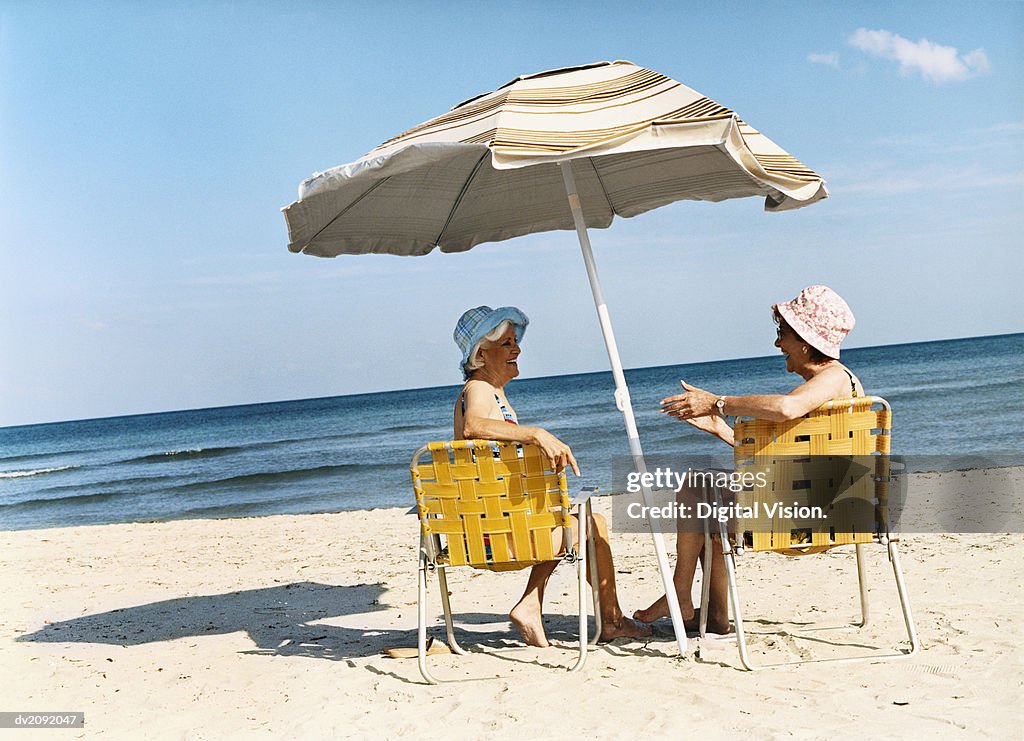 Two Senior Women Sit on Deckchairs Under a Parasol on the Beach, Talking