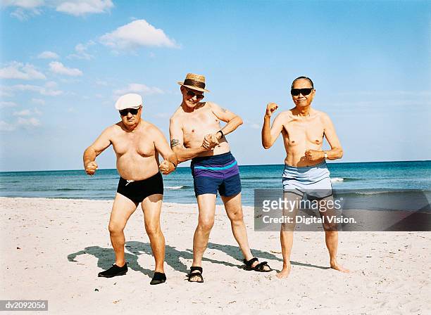 three senior men in swimming trunks stand on the beach flexing their muscles - alardear fotografías e imágenes de stock