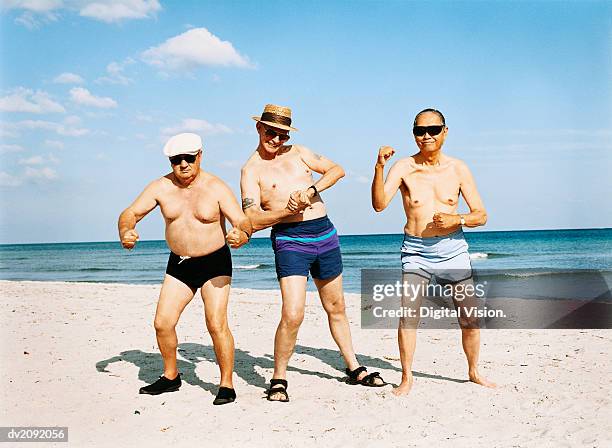 three senior men in swimming trunks stand on the beach flexing their muscles - showing off foto e immagini stock