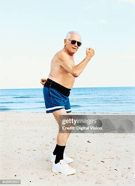 senior man in swimming trunks stands on the beach flexing his muscles - mens swimwear stock pictures, royalty-free photos & images