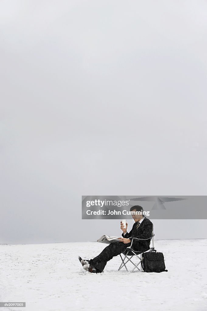 Man Sitting on a Chair Outdoors in Snow and Reading a Newspaper