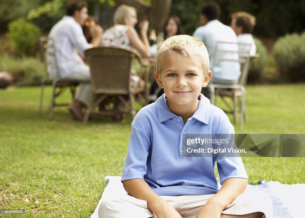 Portrait of a Young Boy Sitting on the Grass in the Garden, People at a Table in the Background