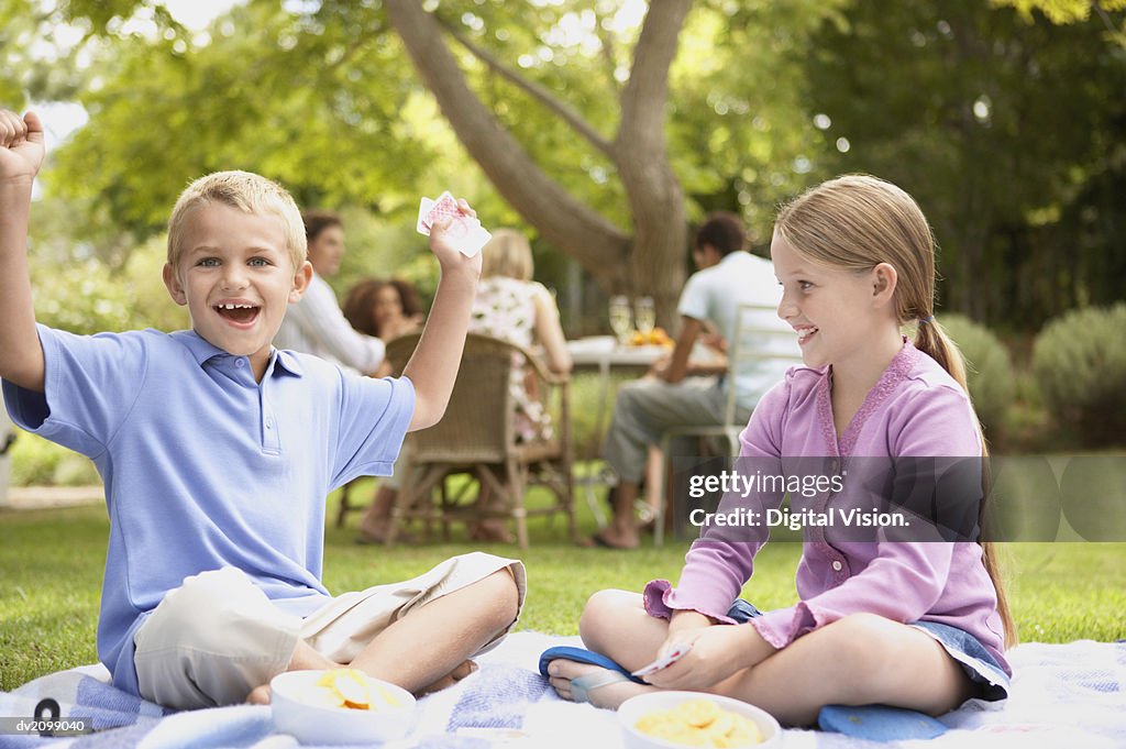 Young Boy and Girl Sit in Their Garden Playing Cards, the Boy Cheering as He Wins the Card Game