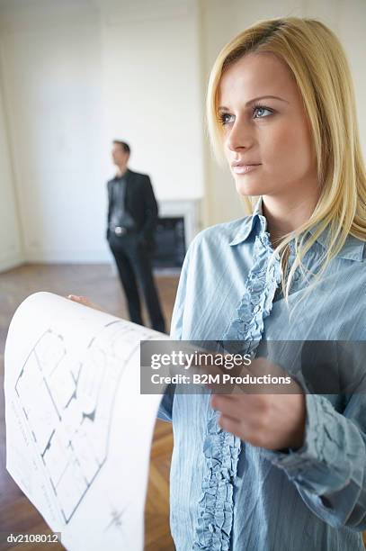 woman holding blueprints in an empty room, with a man standing in the background - shirt met ruches stockfoto's en -beelden