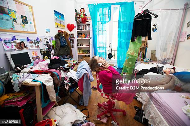 young woman sits on a chair in a messy bedroom using a mobile phone - casual room stock-fotos und bilder