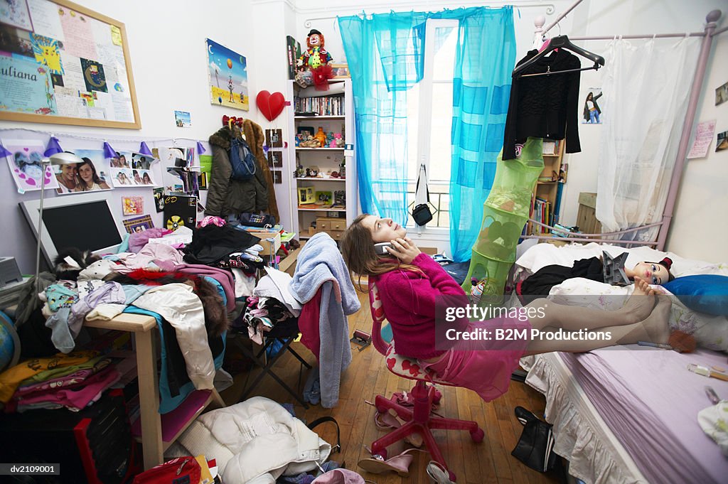 Young Woman Sits on a Chair in a Messy Bedroom Using a Mobile Phone