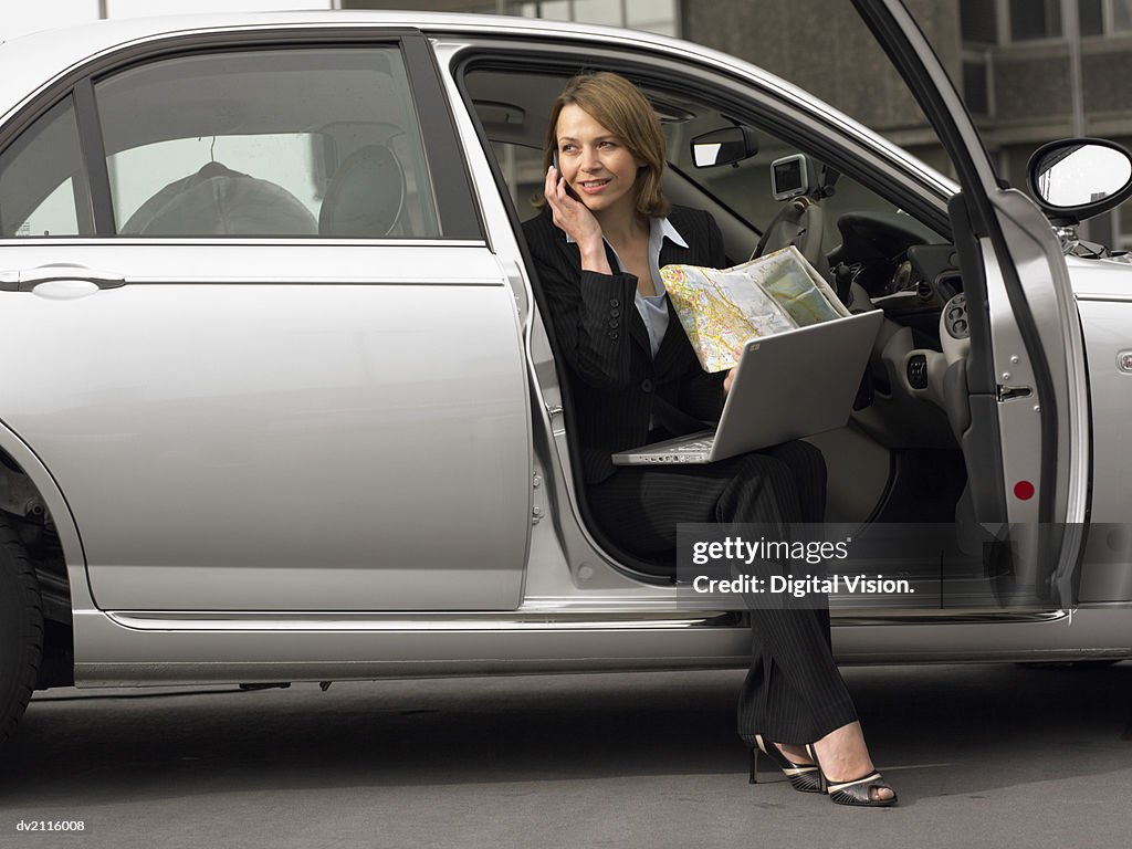 Businesswoman Sitting in Her Car Using a Mobile Phone With a Laptop and a Map