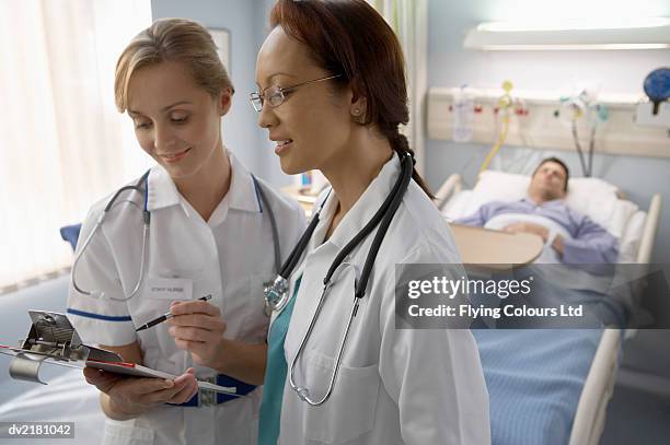 female nurse and doctor stand by a hospital bed discussing the patient's charts - clipboard and glasses imagens e fotografias de stock