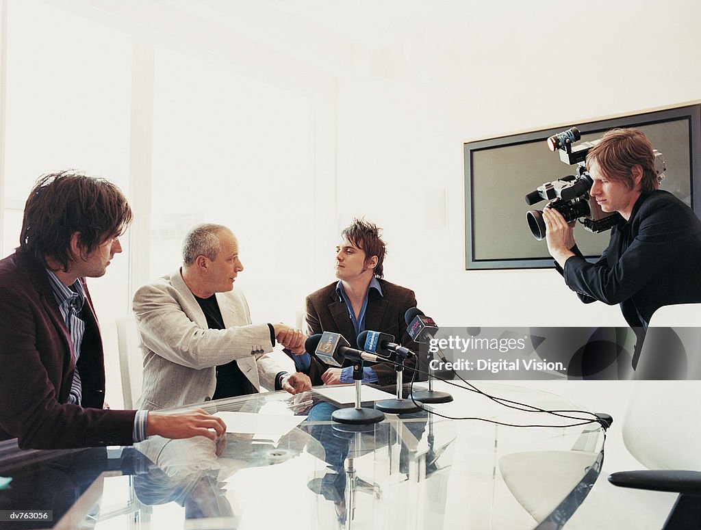Pop Musician Shaking Hands With a Businessman at a News Conference With Microphones and a TV Cameraman
