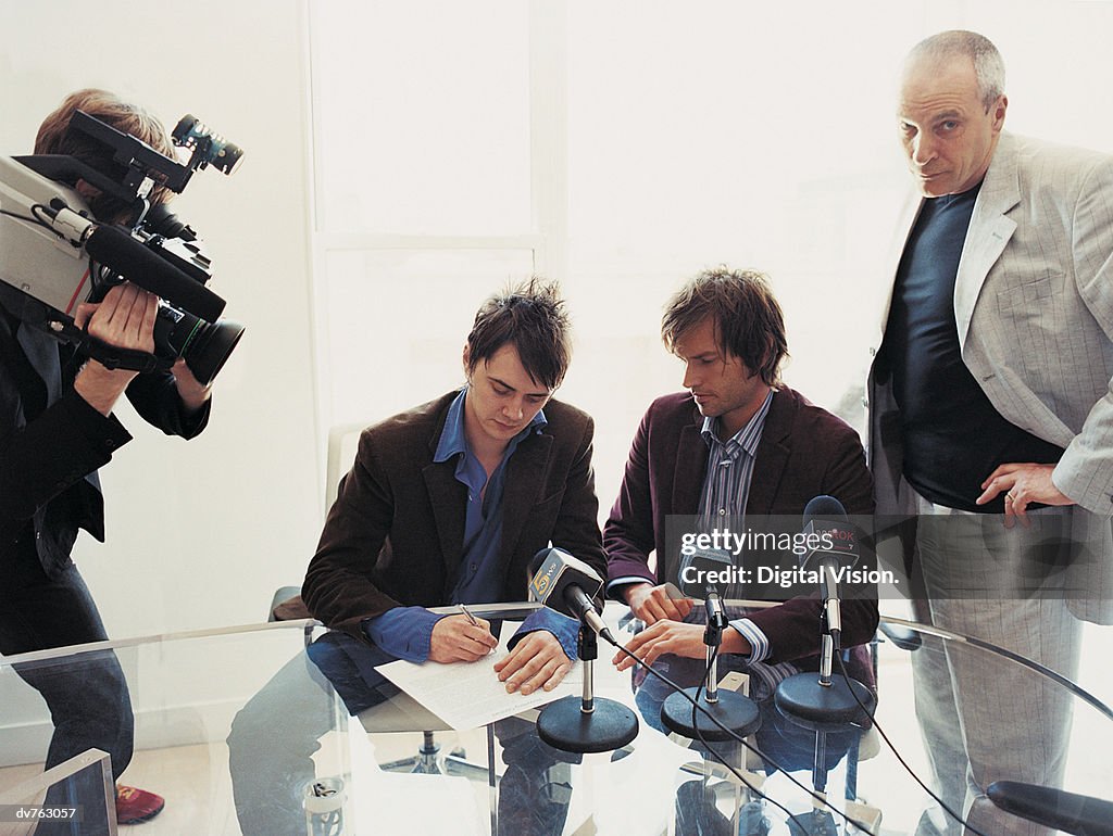 Pop Musicians Signing a Contract in a Conference Room Attended by Their Manager and a TV Cameraman