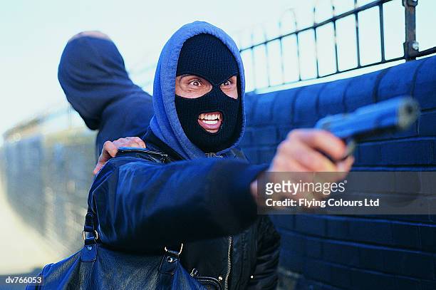male criminal with another man wearing a baraclava aiming a gun - stranger danger stock pictures, royalty-free photos & images