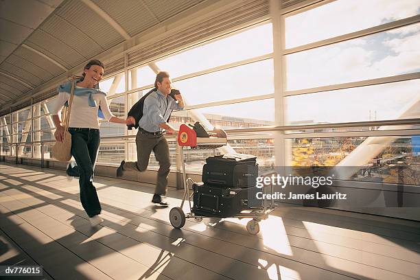 couple rushing down a corridor at the airport, pushing a baggage trolley - luggage trolley stockfoto's en -beelden