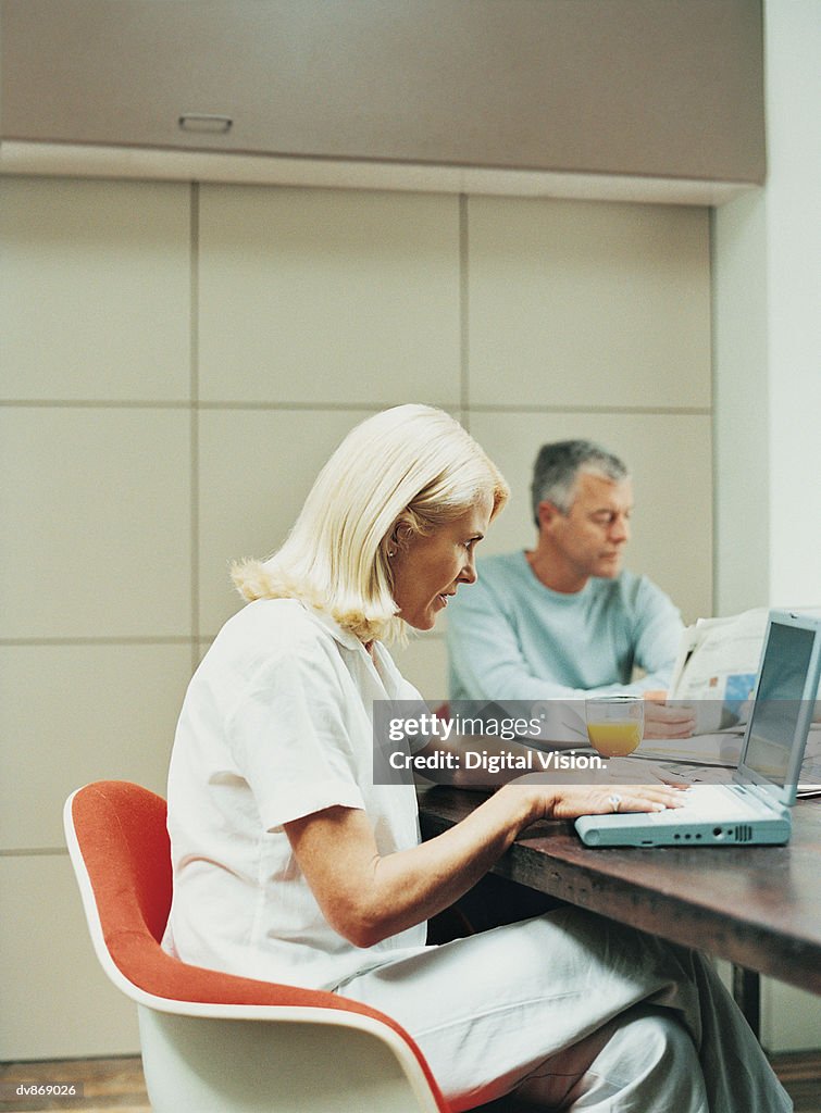 Woman Using a Laptop with a Man Reading a Newspaper