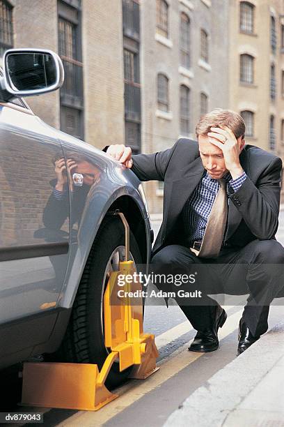 businessman crouching on the kerb looking down at his wheel-clamped car - kerb stock-fotos und bilder