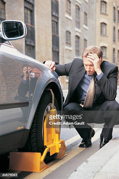 businessman crouching on the kerb looking down at his wheel-clamped car - kerb fotografías e imágenes de stock