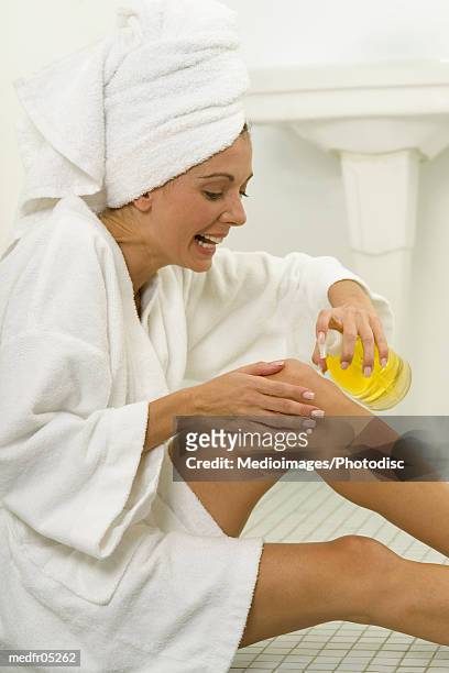 a young woman sitting in a bath robe applying lotion on her legs - bath robe stockfoto's en -beelden