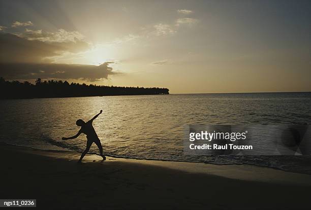 a child, silhouetted by the sunset, throws a rock into the surf. - greater antilles stock-fotos und bilder