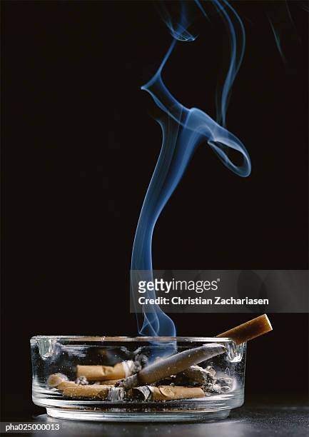 ashtray with cigarette butts and lit cigarette emitting smoke against black background - ashtray stock pictures, royalty-free photos & images