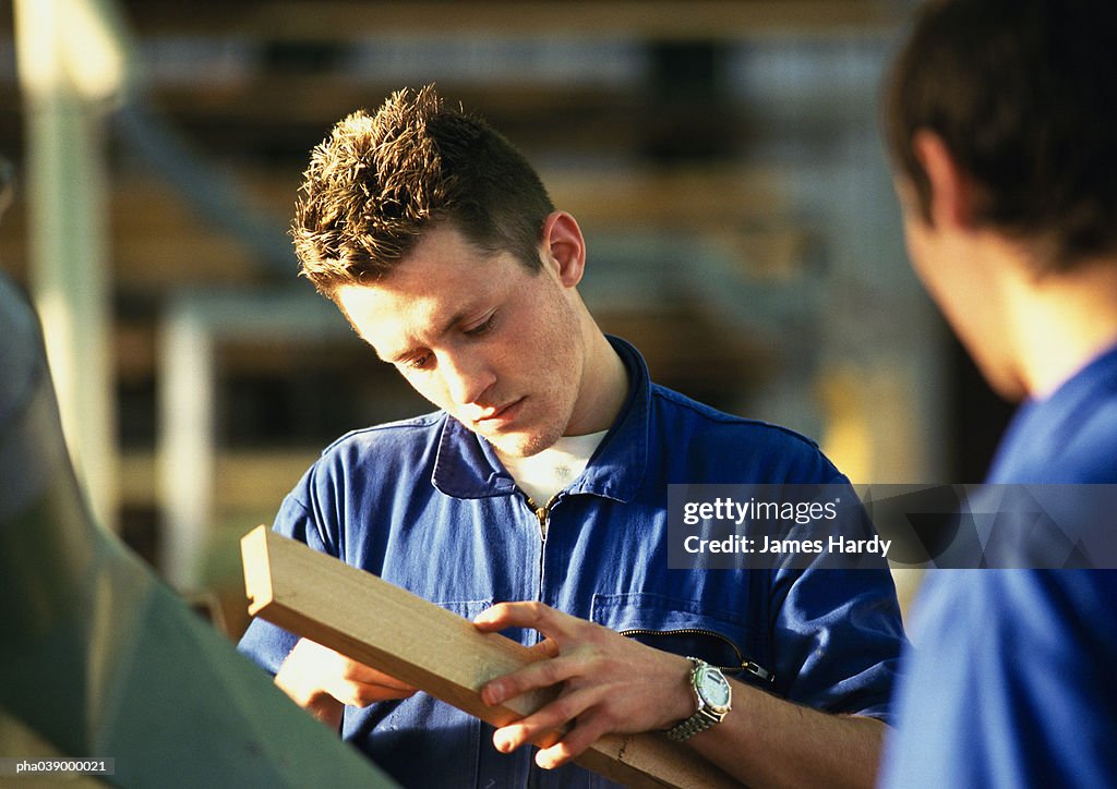 Man in blue coveralls holding plank of wood, second man rear view, blurred in foreground