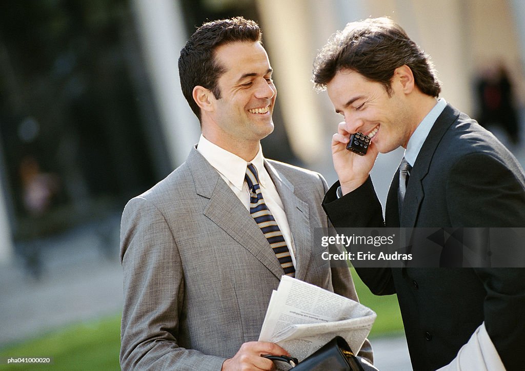 Two businessmen side by side outside, one using cell phone