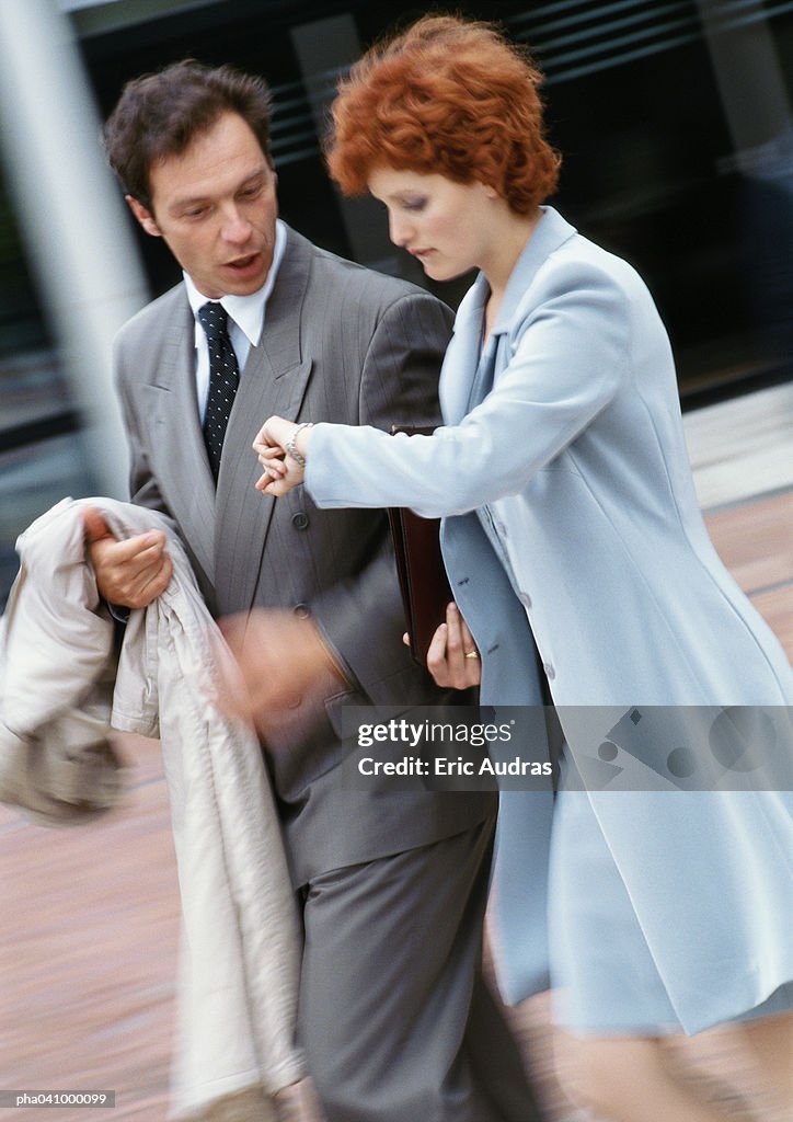 Businessman and businesswoman walking side by side outside, businessman looking at watch