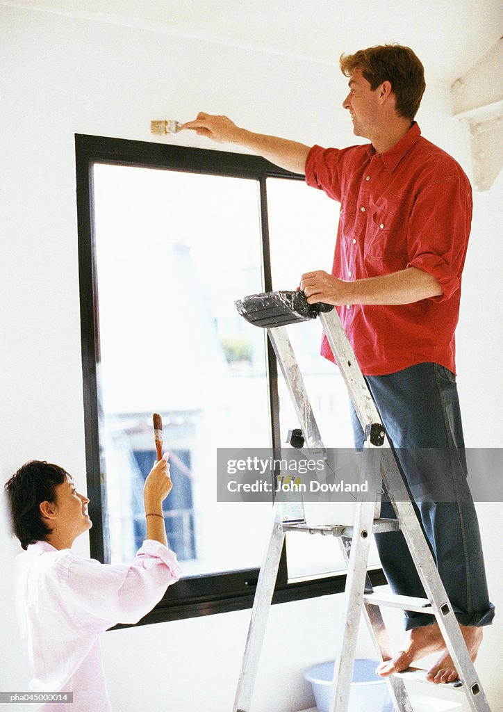 Man standing on ladder, painting wall, woman holding up brush