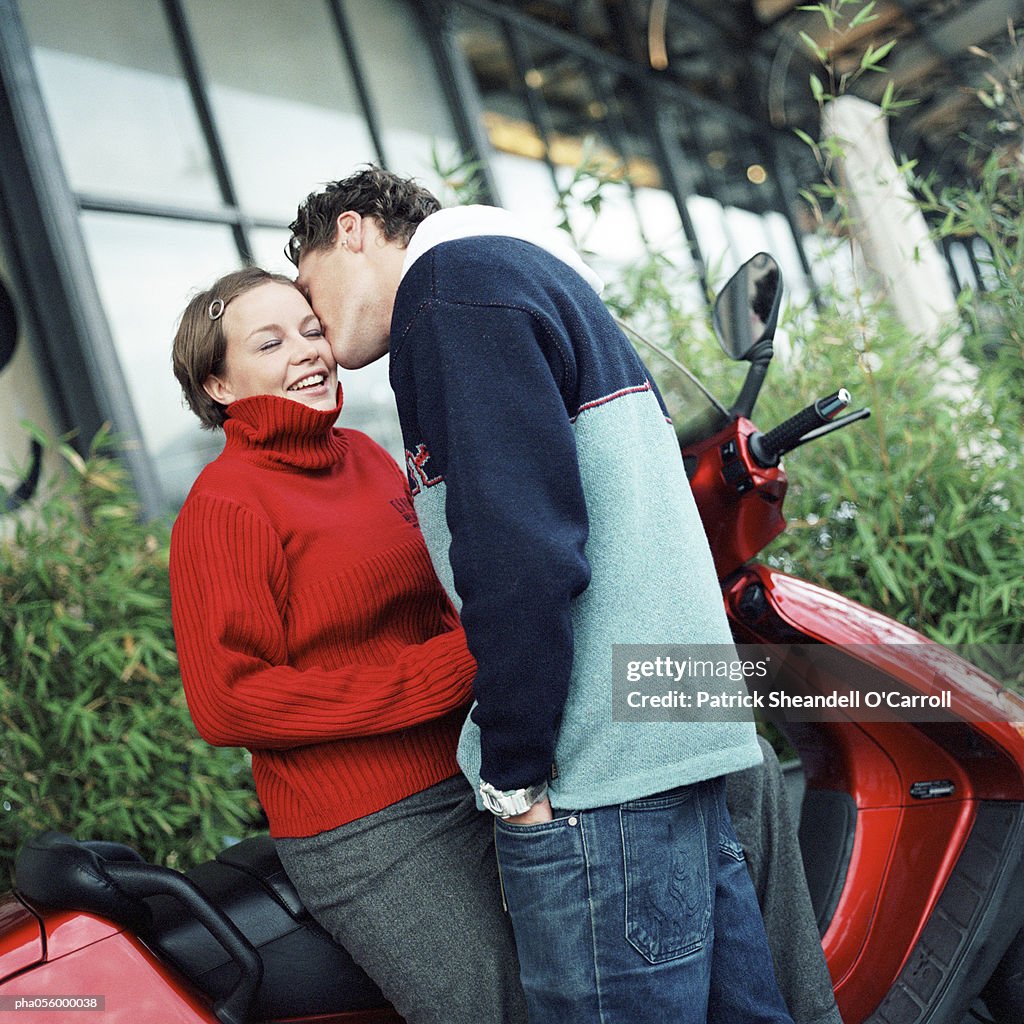 Young man kissing young woman next to scooter