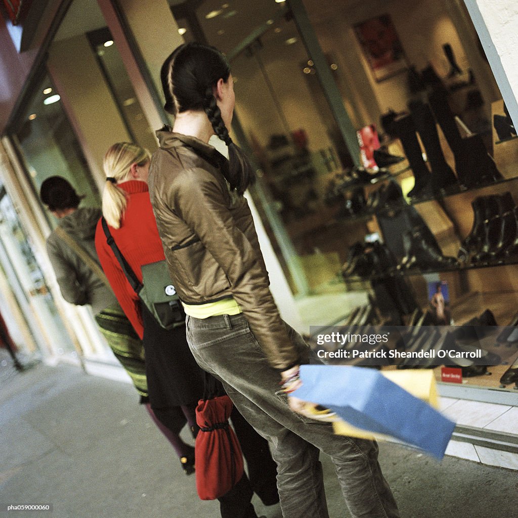 Young woman standing with shopping bag in front of shop window, side view, two other people in background