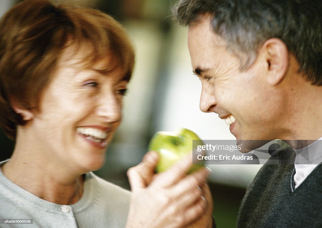 Man and woman smiling at each other, holding apple between them, close up, blurred