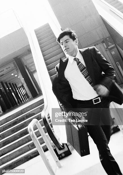 businessman walking away from escalator, holding briefcase, blurred motion, b&w. - necktie run stockfoto's en -beelden