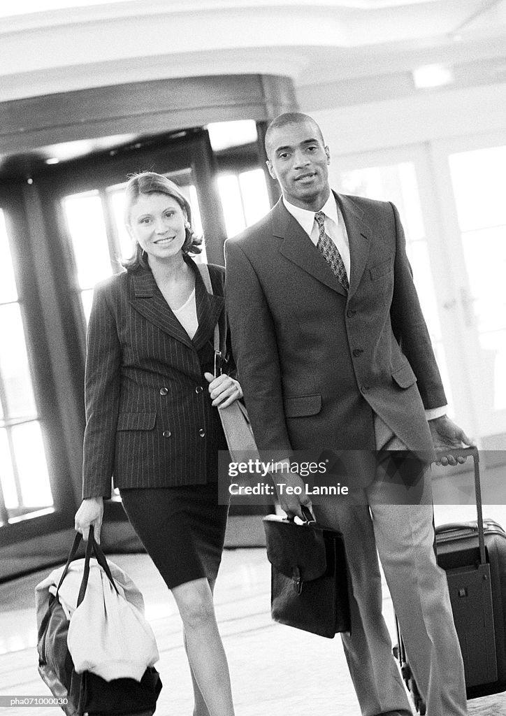 Businessman and businesswoman walking together inside building with luggage, b&w.