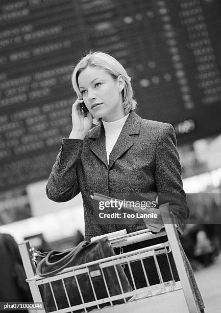 businesswoman on cell phone with luggage caddy in front of arrival and departure board, b&w. - departure board front on fotografías e imágenes de stock