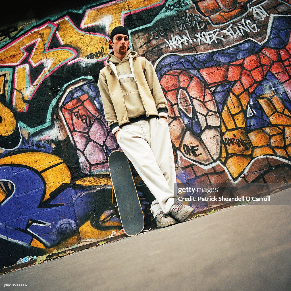 Man with skateboard leaning against graffiti wall, portrait