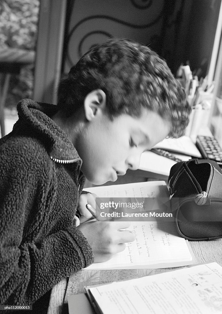 Boy sitting at desk, writing in notebook, side view, b&w