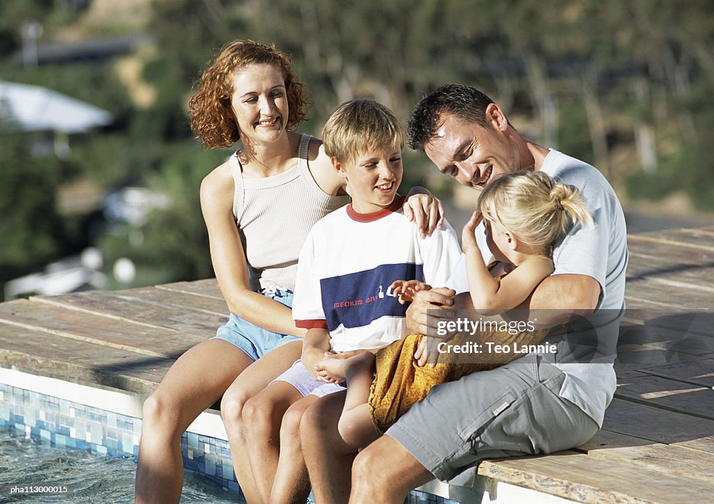 Young family, dressed, sitting on edge of pool.